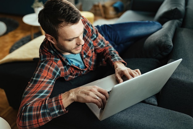 Jeune homme surfant sur le net sur un ordinateur tout en se relaxant dans le salon