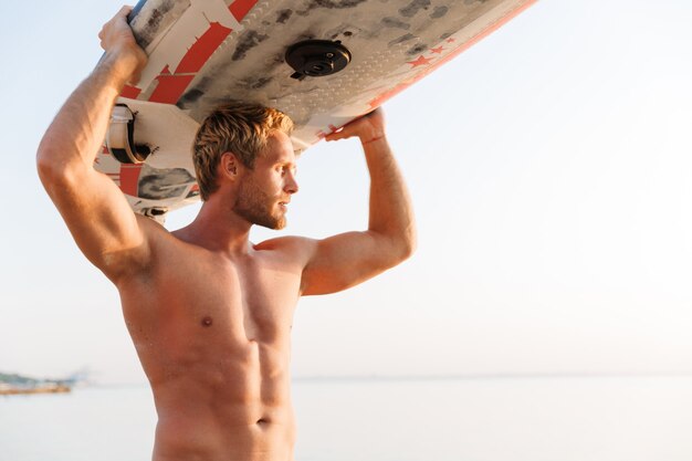 Jeune homme surfeur torse nu masculin portant sa planche de surf par océan au lever du soleil