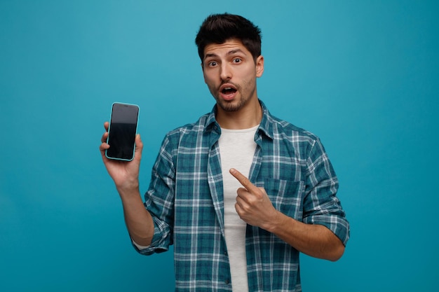 Jeune homme surpris montrant un téléphone portable à la caméra en le pointant en regardant la caméra isolée sur fond bleu