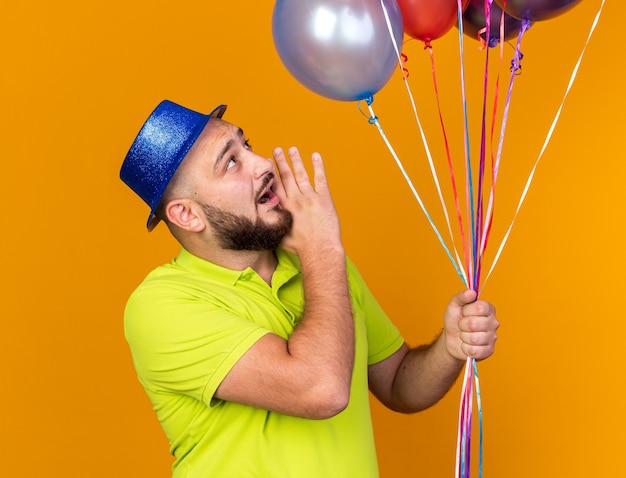 Jeune homme surpris portant un chapeau de fête tenant et regardant des ballons appelant quelqu'un