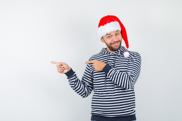 Jeune homme en sweat à capuche, bonnet de noel pointant vers la gauche et à la recherche d'hésitation ,