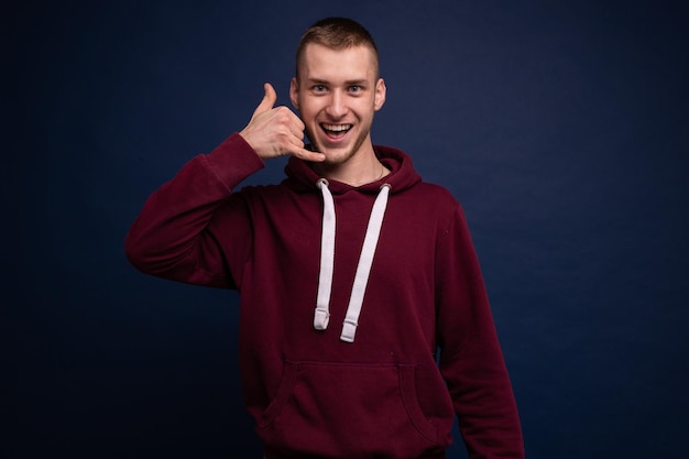 Jeune homme en sweat à capuche rouge et jeans posant sur fond bleu et montrant le geste d'appel