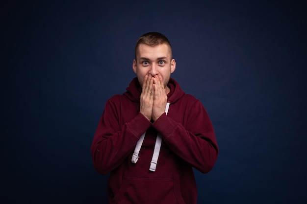 Jeune homme en sweat à capuche rouge et jeans posant sur fond bleu surpris et couvre son visage avec ses mains