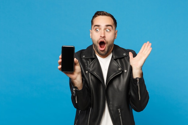 Jeune homme en t-shirt blanc veste noire tenir un téléphone portable avec un écran vide vierge pour le contenu promotionnel isolé sur fond de mur bleu portrait en studio. Concept de mode de vie des gens. Maquette de l'espace de copie