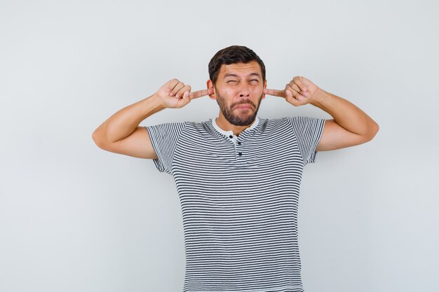 Jeune homme en t-shirt bouchant les oreilles avec les doigts et ayant l'air irrité, vue de face.