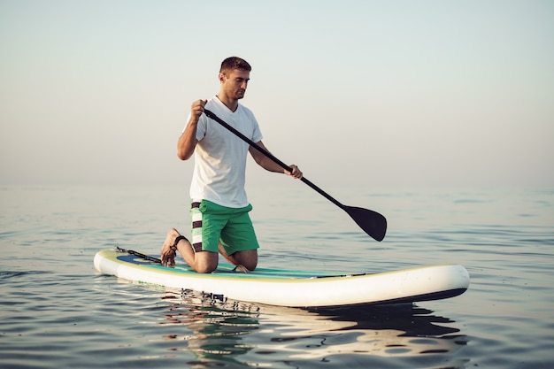 Jeune homme en t-shirt et short flottant sur planche de SUP en mer