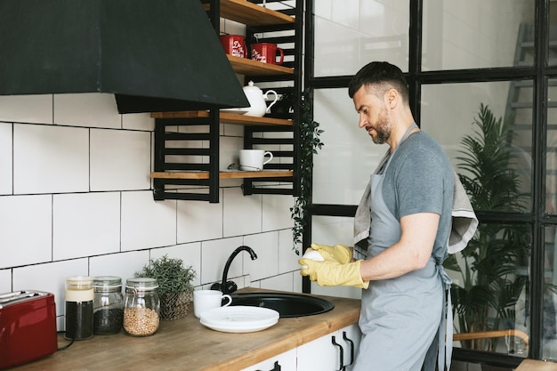 jeune homme en tablier et gants fait les tâches ménagères lave la vaisselle avec une serviette sur son épaule hommes tâches ménagères aide ménagère dans une cuisine élégante dans un appartement moderne