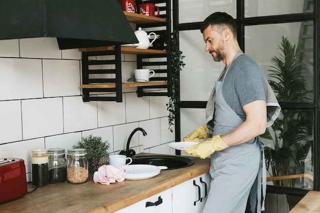 jeune homme en tablier et gants fait les tâches ménagères lave la vaisselle avec une serviette sur son épaule hommes tâches ménagères aide ménagère dans une cuisine élégante dans un appartement moderne