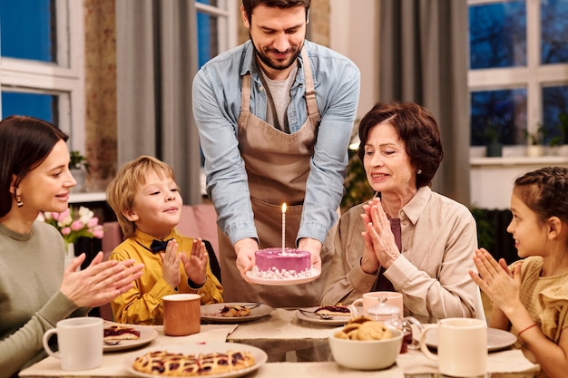 Jeune homme en tablier mettant une assiette avec un gâteau glacé fait maison avec une bougie allumée sur une table servie avant de dîner festif en famille