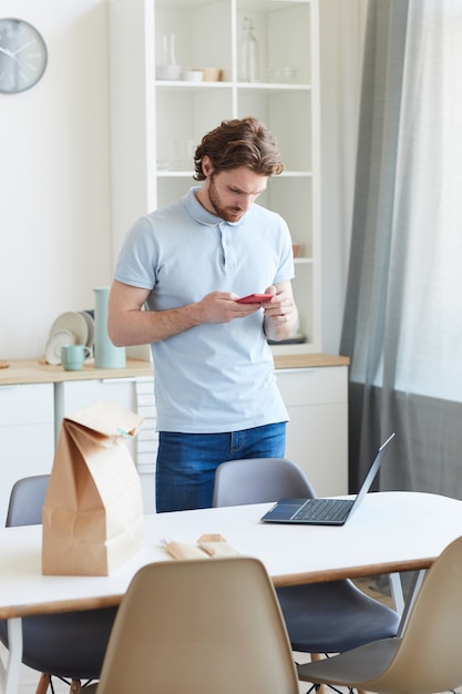 Jeune homme tapant un message sur téléphone mobile en se tenant debout dans la cuisine à la maison