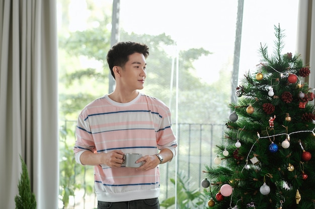 Jeune homme avec une tasse de café comme cadeau de vacances avec un arbre décoré en arrière-plan