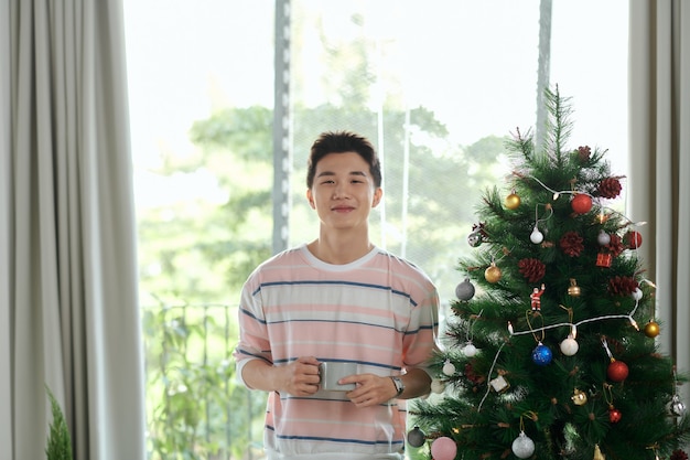 Jeune homme avec une tasse de café comme cadeau de vacances avec un arbre décoré en arrière-plan