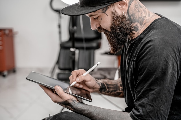 Jeune homme tatoueur avec barbe tenant un crayon et un croquis semblant positif et heureux debout et souriant dans le lieu de l'atelier.