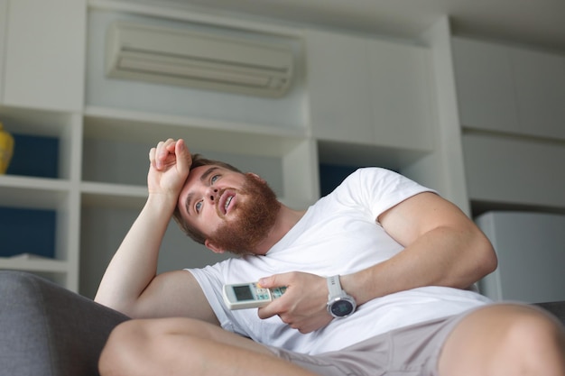 Jeune homme avec télécommande de climatiseur utilisant le climatiseur à la maison, le gars se rafraîchit