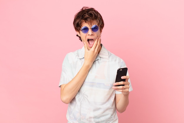 Jeune homme avec un téléphone portable avec la bouche et les yeux grands ouverts et la main sur le menton