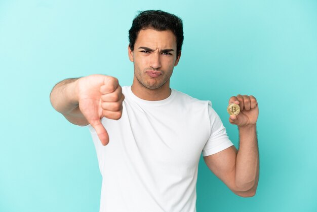 Jeune homme tenant un Bitcoin sur fond bleu isolé montrant le pouce vers le bas avec une expression négative