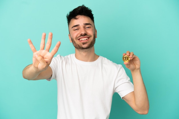 Jeune homme tenant un Bitcoin isolé sur fond bleu heureux et comptant quatre avec les doigts