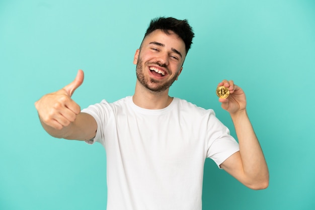 Jeune homme tenant un Bitcoin isolé sur fond bleu avec les pouces vers le haut parce que quelque chose de bien s'est produit