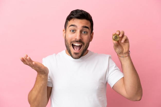 Jeune homme tenant un Bitcoin isolé sur fond rose avec une expression faciale choquée