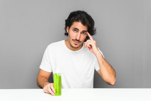 Jeune homme tenant une bouteille d'aloe vera pointant sa tempe avec le doigt, pensant, concentré sur une tâche.