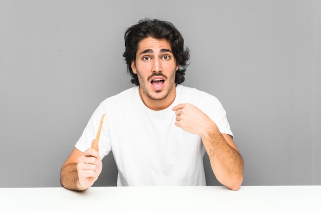 Jeune homme tenant une brosse à dents surpris de se pointer du doigt, souriant largement.