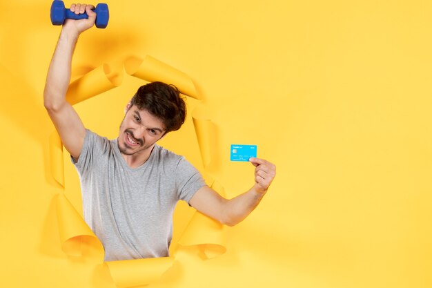 Jeune homme tenant une carte de crédit et un haltère sur fond jaune, des séances d'entraînement adaptées à l'argent de l'athlète