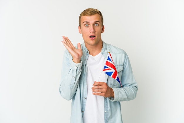 Jeune homme tenant un drapeau du Royaume-Uni isolé sur un mur blanc surpris et choqué