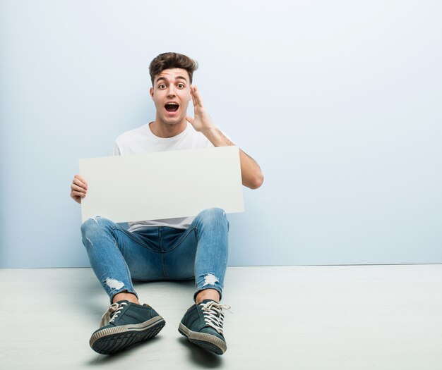 Jeune homme tenant une pancarte assise sur son plancher de maison criant excité à l'avant.