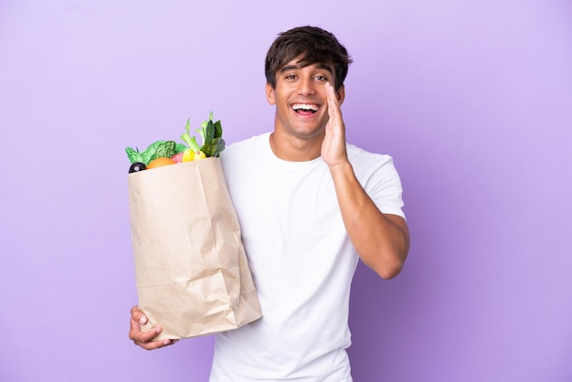 Jeune homme tenant un sac d'épicerie isolé sur fond violet criant avec la bouche grande ouverte