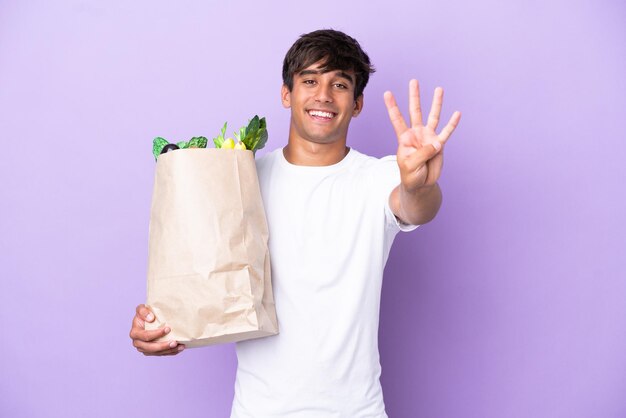 Jeune homme tenant un sac d'épicerie isolé sur fond violet heureux et comptant quatre avec les doigts