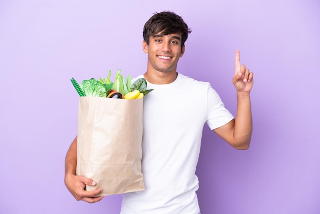 Jeune homme tenant un sac d'épicerie isolé sur fond violet pointant vers le haut une excellente idée