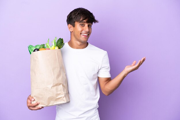 Jeune homme tenant un sac d'épicerie isolé sur fond violet tendant les mains sur le côté pour inviter à venir