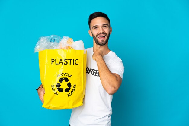 Jeune homme tenant un sac plein de bouteilles en plastique à recycler isolé sur mur bleu célébrant une victoire