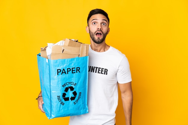 Jeune homme tenant un sac de recyclage plein de papier à recycler isolé sur un mur blanc en levant et avec une expression surprise