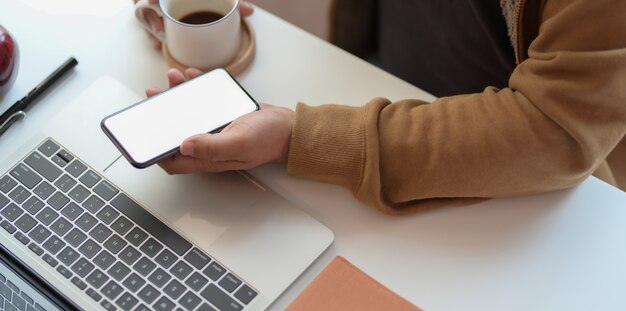 Jeune homme tenant un smartphone écran vide tout en travaillant sur un ordinateur portable et de boire une tasse de café