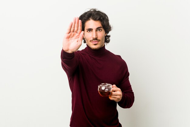 Jeune homme tenant une tasse de thé debout avec la main tendue montrant le panneau d'arrêt, vous empêchant.