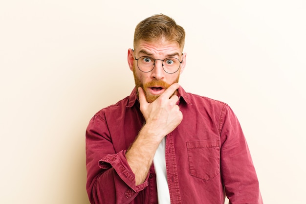 Jeune homme à tête rouge avec la bouche et les yeux grands ouverts et la main sur le menton, se sentant désagréablement choqué, disant quoi ou wow sur fond beige