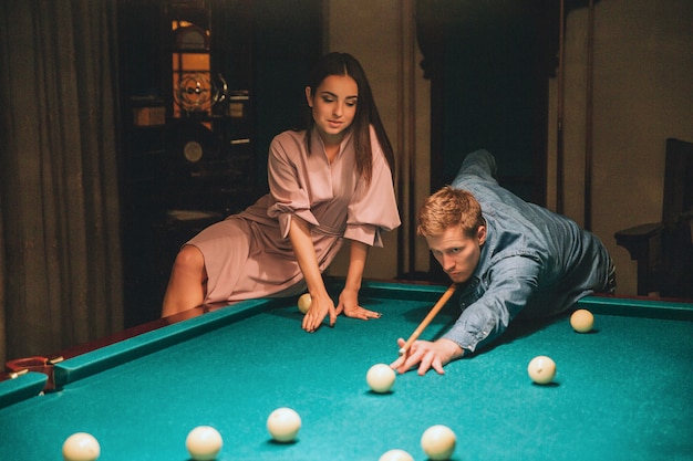 Jeune homme tête rouge jouer au jeu de billard. Il a l'air concentré. Beau modèle assis au coin de la table et appuyé dessus. Ils sont à l'intérieur.