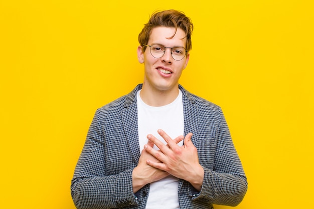 Jeune homme à la tête rouge se sentant romantique, heureux et amoureux, souriant gaiement et tenant les mains proches du cœur contre orange