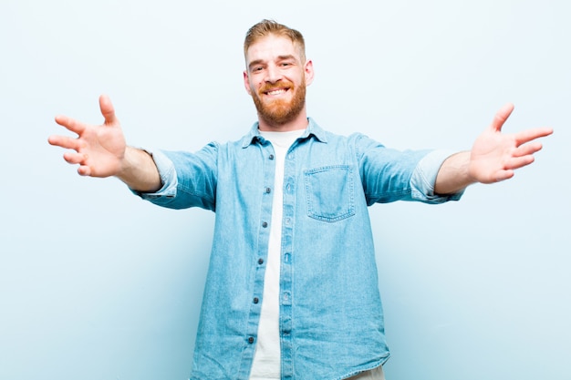 Jeune homme à tête rouge souriant joyeusement donnant un câlin de bienvenue chaleureux, amical et affectueux, se sentant heureux et adorable sur un mur bleu doux