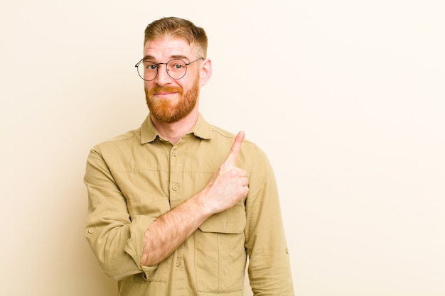 Jeune homme tête rouge souriant joyeusement, se sentant heureux et pointant vers le côté et vers le haut, montrant l'objet dans l'espace de copie sur le mur beige