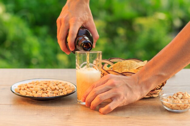 Le jeune homme tient une bouteille de bière et remplit le verre. Main masculine versant de la bière dans du verre sur une table en bois avec des croustilles dans un panier en osier, des arachides dans une assiette et un bol