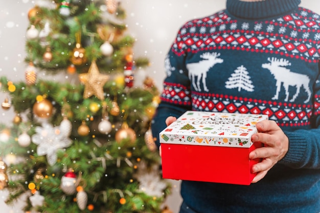 Un jeune homme tient un cadeau de Noël à l'appareil photo sur fond d'arbre décoré