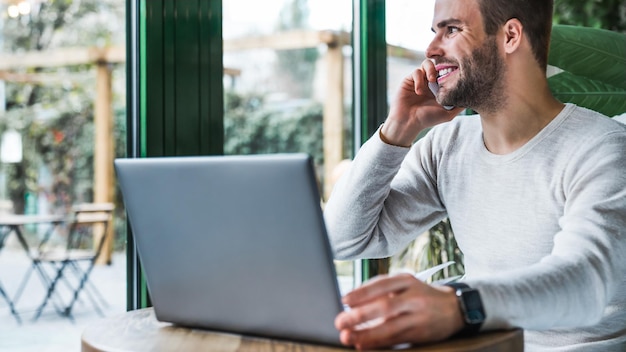 Jeune homme travaillant dans un café utilisant un téléphone portable et un ordinateur portable tout en regardant par la fenêtre