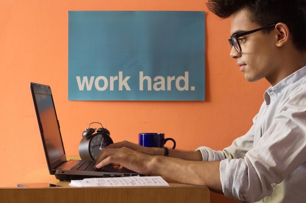 Photo un jeune homme travaillant dur à domicile sur un ordinateur portable placé sur un bureau à côté d'une affiche de travail dur sur un mur