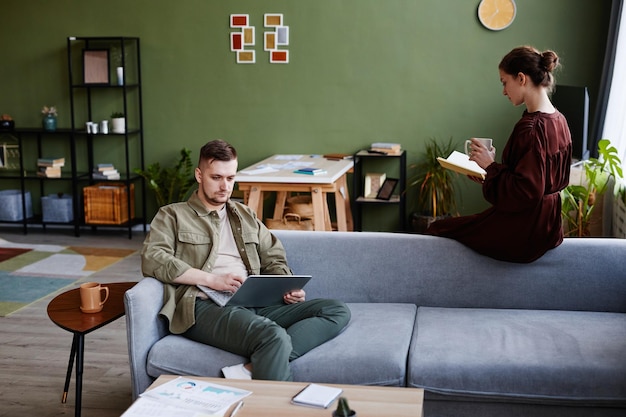 Jeune homme travaillant en ligne sur un ordinateur portable assis sur un canapé avec une femme lisant un livre et buvant du café