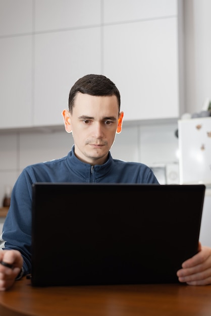 Un jeune homme travaillant en ligne avec un ordinateur portable assis seul à une table à l'intérieur de son appartement moderne