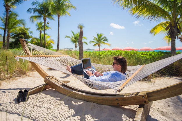 Jeune homme travaillant sur un ordinateur portable dans un hamac sur une plage tropicale