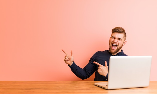 Jeune homme travaillant avec son ordinateur portable pointant avec des index sur un espace de copie, exprimant son enthousiasme et son désir.