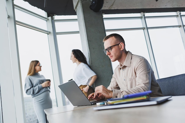 Un jeune homme travaille dans le bureau sur un ordinateur portable tout en étant assis à la table contre le fond des collègues espace de travail moderne avec les employés de bureau